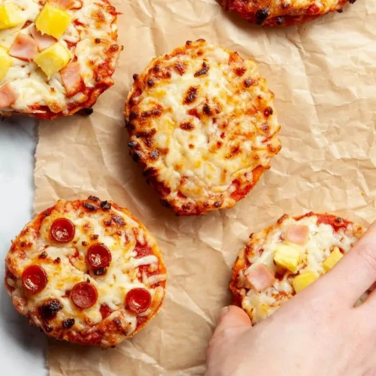 This image is a set of 5 mini pizza bites on a crinkled brown paper background. 1 pepperoni, 2 ham and pineapple, and 1 cheese. A hand is grabbing the bottom one and that one was pineapple.