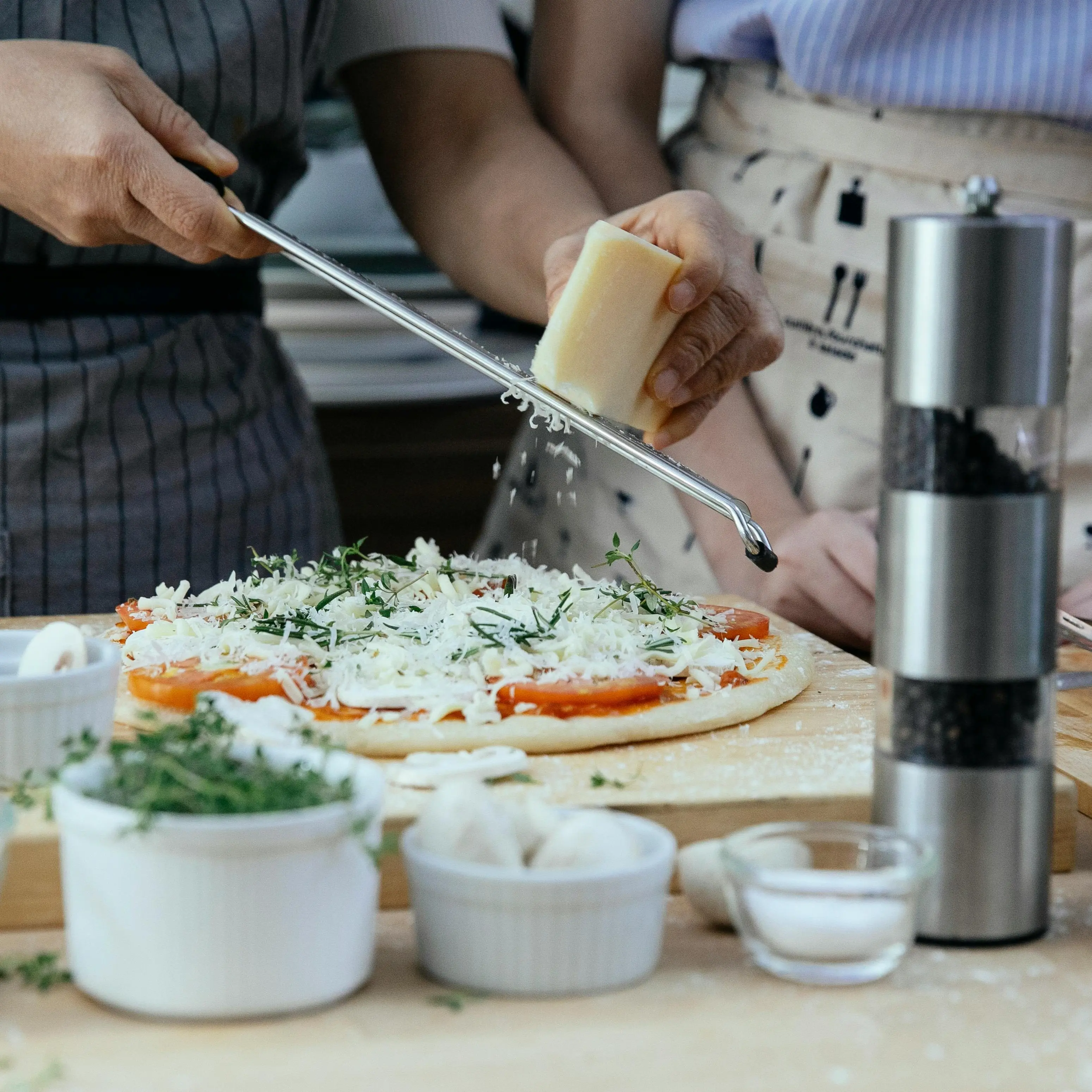 This image is a chef and assistant grating white parmesan cheese on top of a delicious ready to bake dough, while resting on a flour coated cutting board. There are also ingredients in the frame as well. Like tall pepper shaker and a small glass bowl of salt.