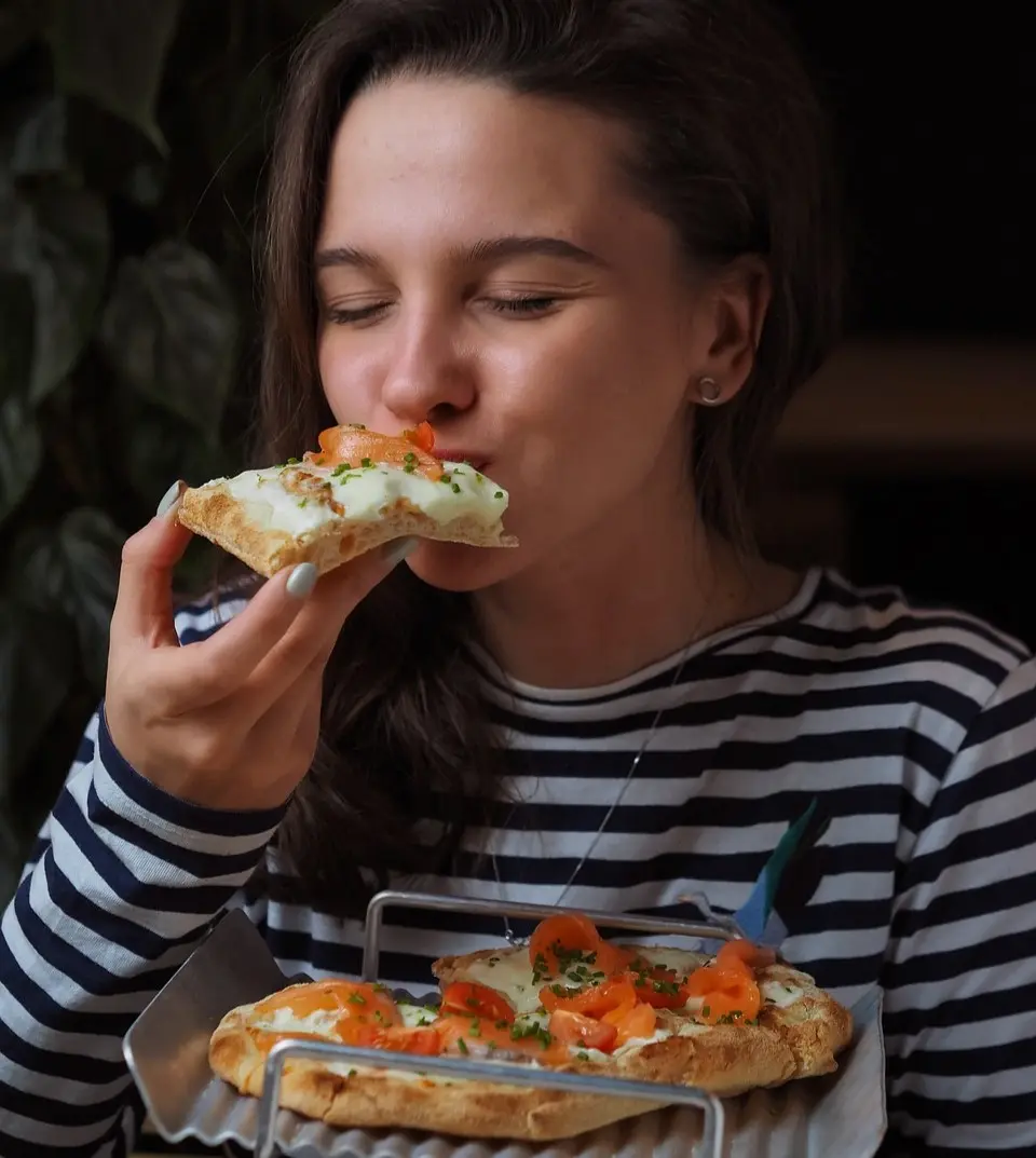 This image is a young girl who is taking her first bite of her mini pizza. She has the slice in one hand and the whole pizza in the other hand.
